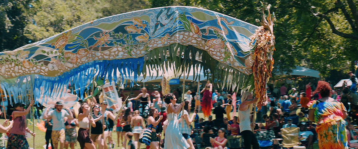 Festivalgoers carry a large, flowing sun dragon puppet across a wooded lawn during a festival.